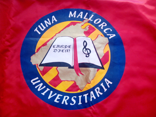 imagen de la bandera de tuna mallorca universitaria con los colores de distrito, el mapa de Mallorca, los colores de nuestra bandera, un libro con la clave de sol y a su izquierda la expresión CARPE DIEM. Es la forma de representar la agrupación que comparte estudiantina Tuna Mallorca